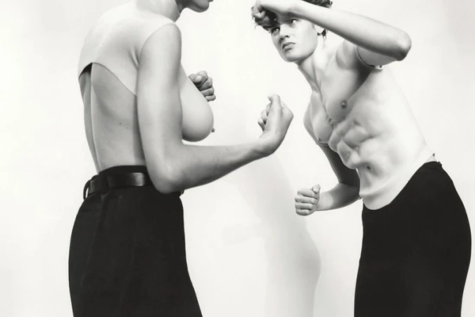 Two shirtless people face each other in a black and white studio shot, with fists raised, wearing black pants against a white background.