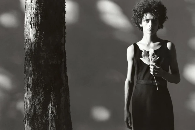 Monochromatic fashion portrait of a person with curly hair in a sleeveless dress, holding a leaf, standing next to a tree trunk.