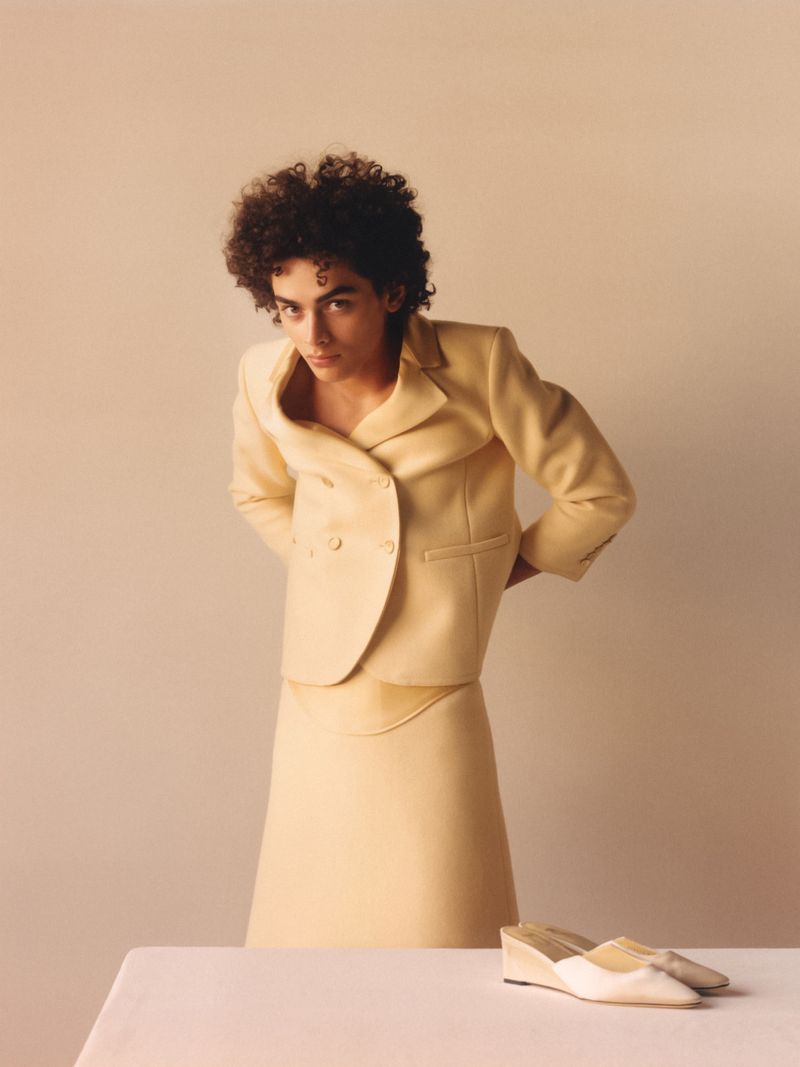 Adult woman with curly hair poses indoors wearing a tailored yellow skirt suit with hands behind her back near a pair of shoes.