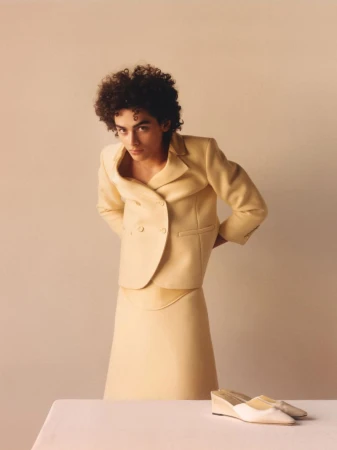 Adult woman with curly hair poses indoors wearing a tailored yellow skirt suit with hands behind her back near a pair of shoes.