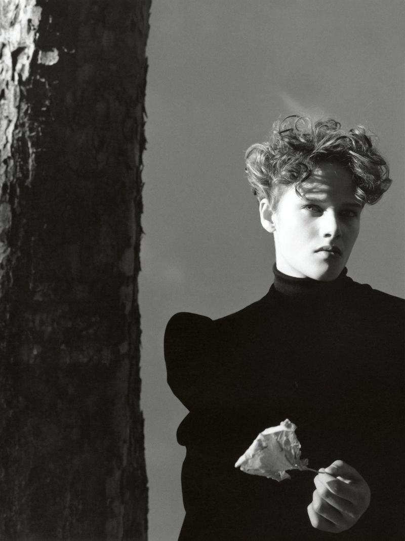 Monochrome portrait of a person with short, curly hair in a dark turtleneck, holding a wilted flower against a stark sky.