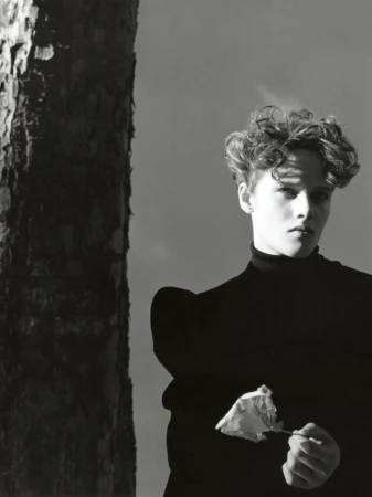 Monochrome portrait of a person with short, curly hair in a dark turtleneck, holding a wilted flower against a stark sky.