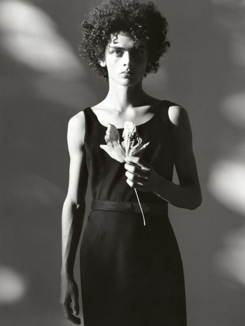 Monochrome studio portrait of a slim adult with curly hair in a dress, holding a dried leaf against a gradient background.