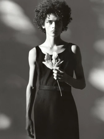 Monochrome studio portrait of a slim adult with curly hair in a dress, holding a dried leaf against a gradient background.