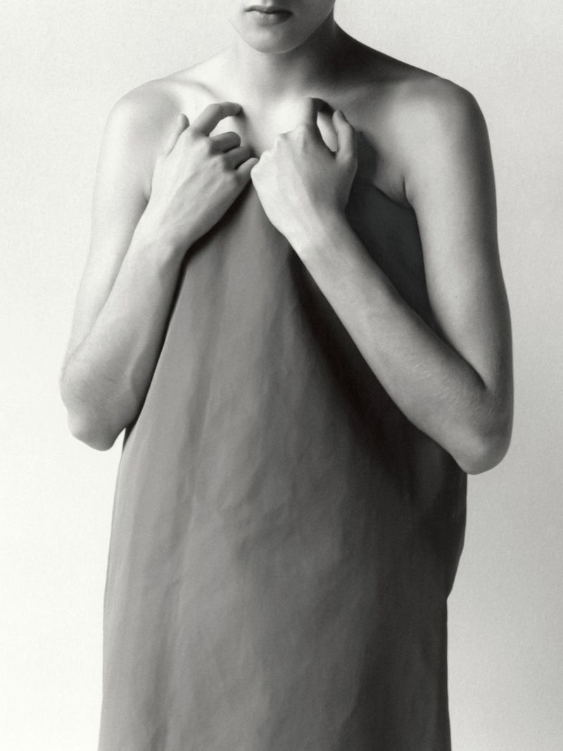 Monochrome studio portrait of an adult with hands holding a draping fabric, emphasizing texture and minimal composition against a plain backdrop.
