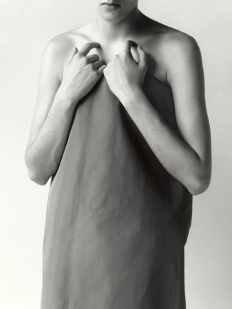 Monochrome studio portrait of an adult with hands holding a draping fabric, emphasizing texture and minimal composition against a plain backdrop.