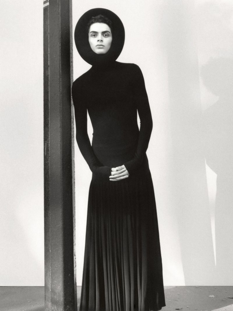 Fashion shot of a person in an all-black outfit featuring a long-sleeved top, a wide-brimmed hat, and a pleated skirt leaning on a pole.