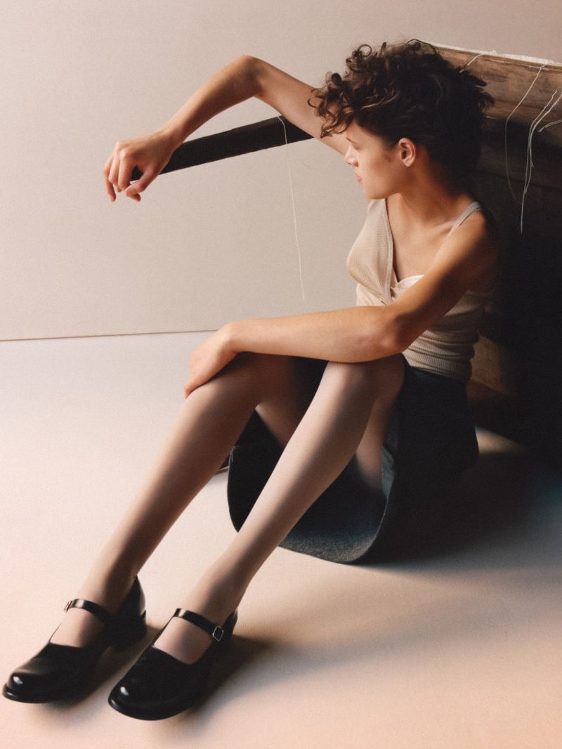 Young woman poses indoors with string, wearing tank top, short skirt, and Mary Jane shoes against white backdrop with natural lighting.
