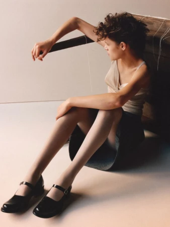 Young woman poses indoors with string, wearing tank top, short skirt, and Mary Jane shoes against white backdrop with natural lighting.