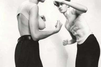 Two models wearing surreal anatomical prosthetic tops facing each other in boxing stances against a plain backdrop.