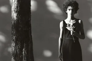 Model with curly hair holding a dried flower in a black dress next to a large tree trunk in a monochrome fashion image.
