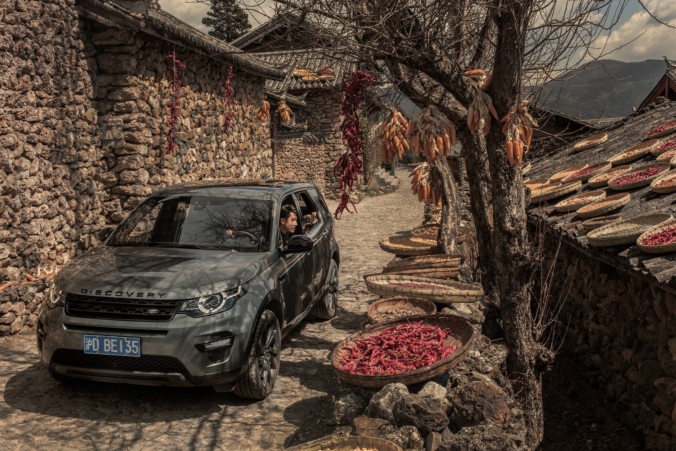 SUV parked on cobblestone street in rural village, China. Wicker baskets with red chilies and corn drying in the sun.