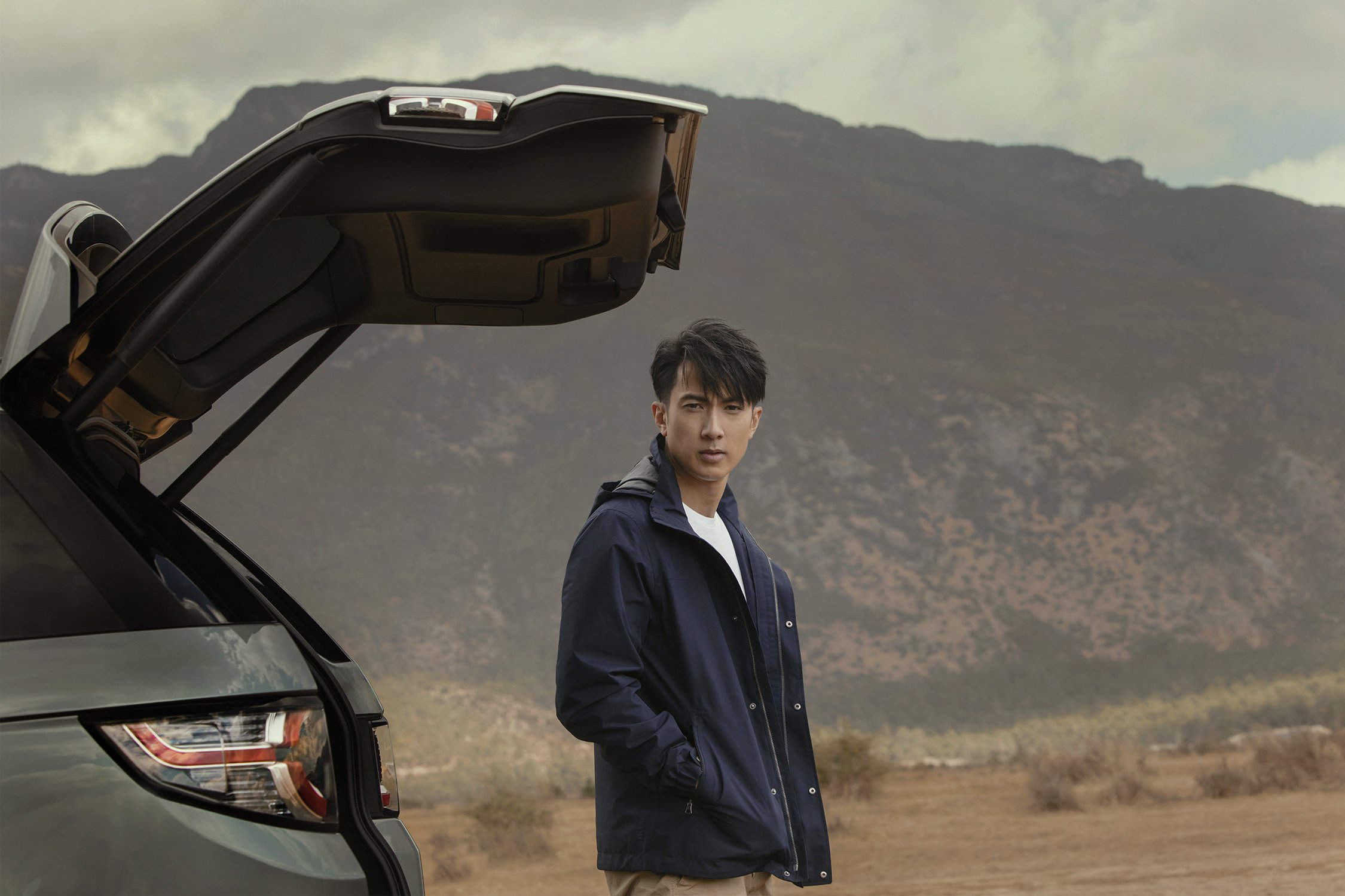 An adult Asian man stands in front of a gray SUV with the trunk open in a scenic outdoor location with mountains in the background.