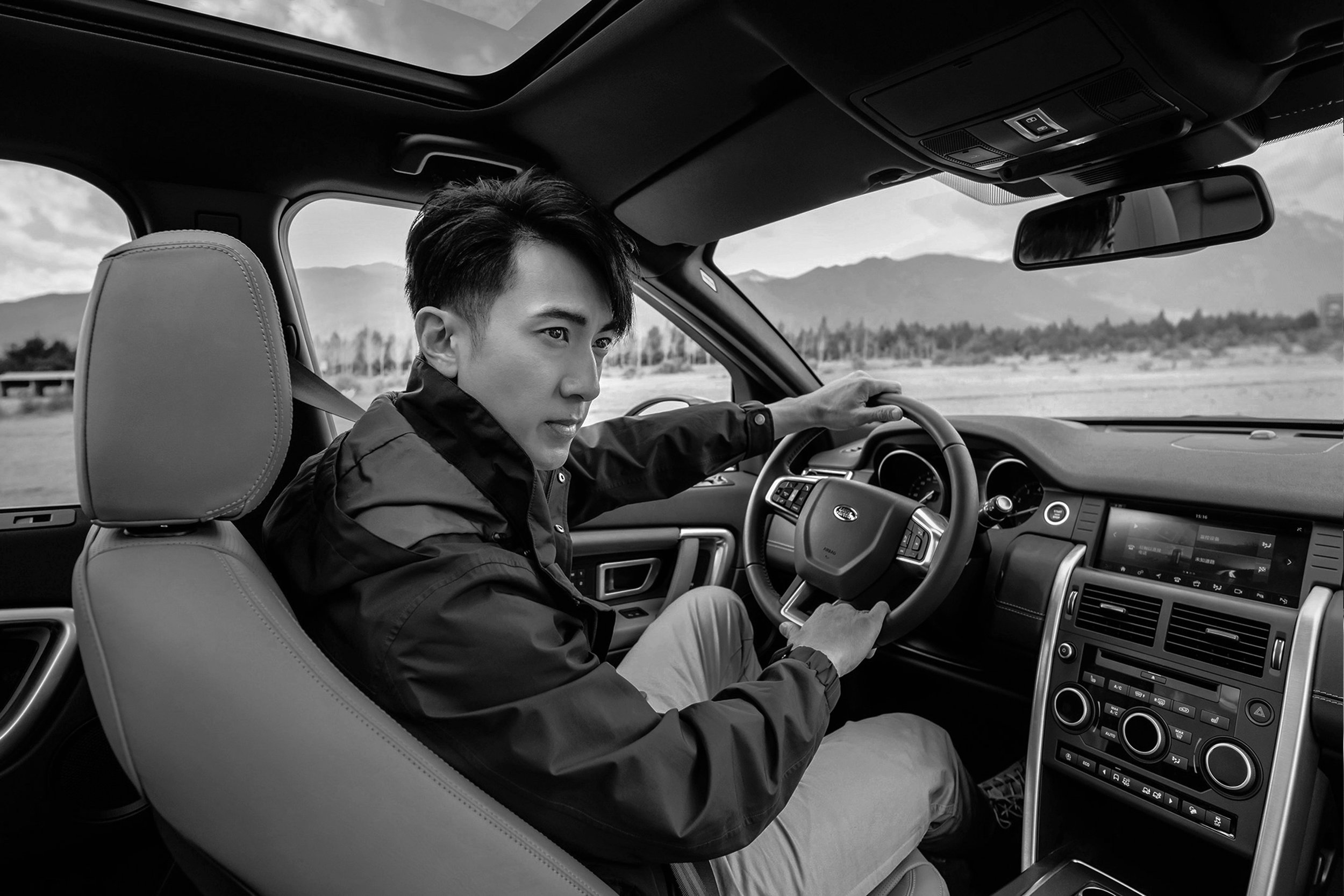 A man sits inside a car with an open sunroof, holding the steering wheel, mountains visible in the background. Automotive shot.