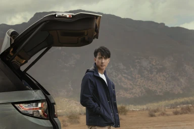 Man in a dark navy blue jacket standing next to an open SUV trunk with mountain terrain in the background
