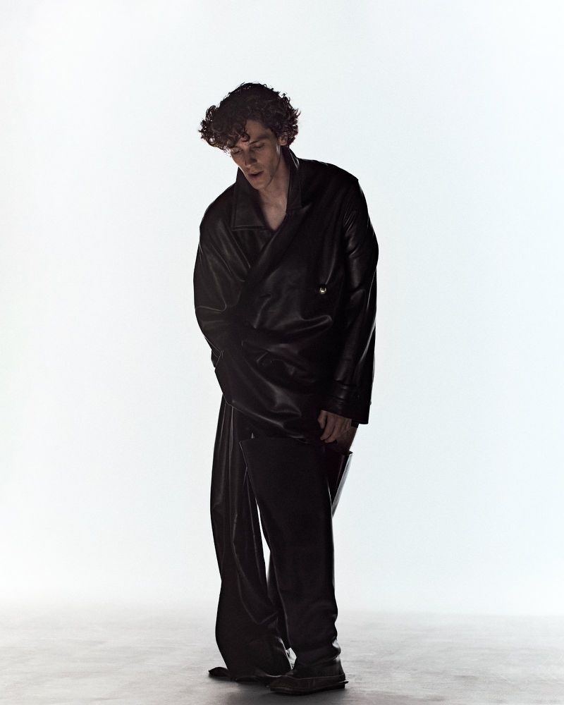 Actor D'Pharaoh Woon-A-Tai standing in an oversized black leather jacket and matching trousers against a bright white backdrop.