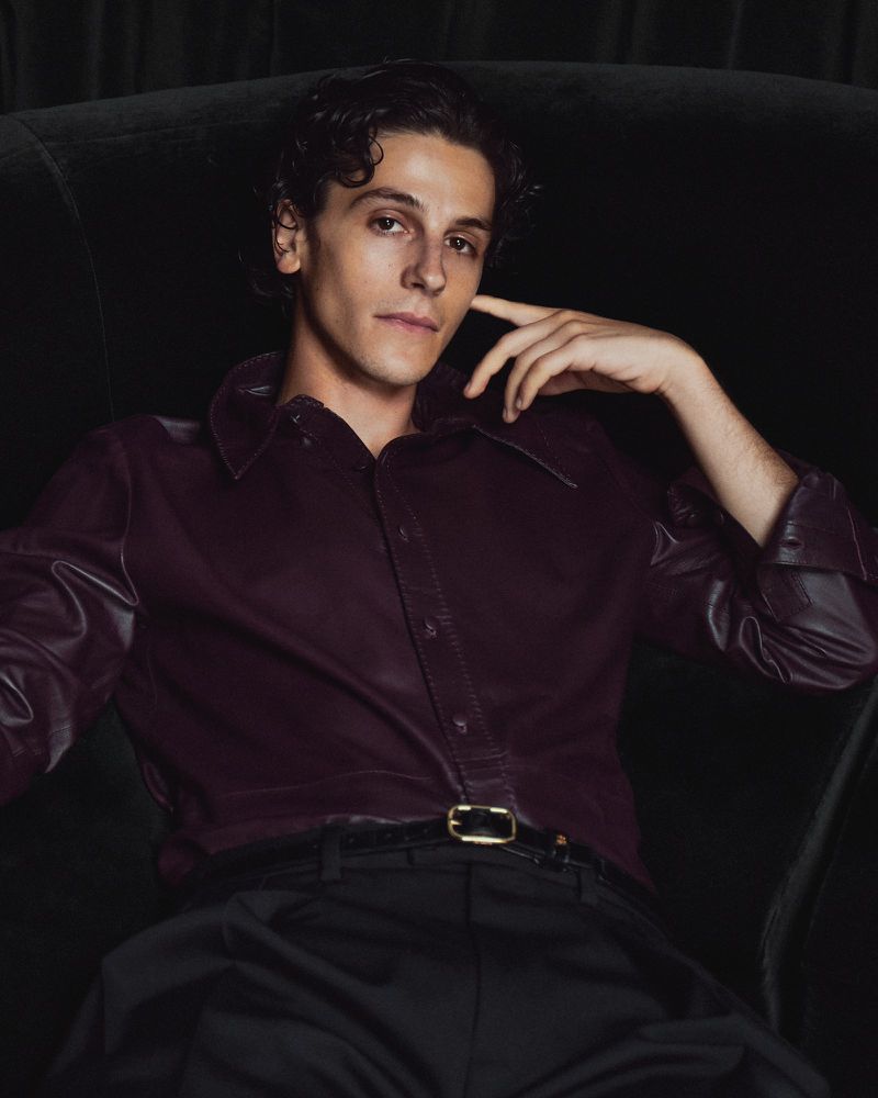 Portrait of a young adult man in a burgundy collared shirt and black pants, seated in a dark green armchair against a dark background.