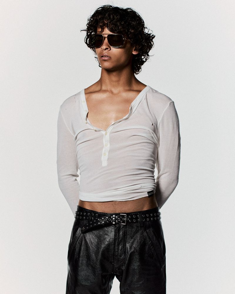 Young adult male with curly hair and sunglasses poses in a white long-sleeved shirt and black leather pants against a plain white backdrop.