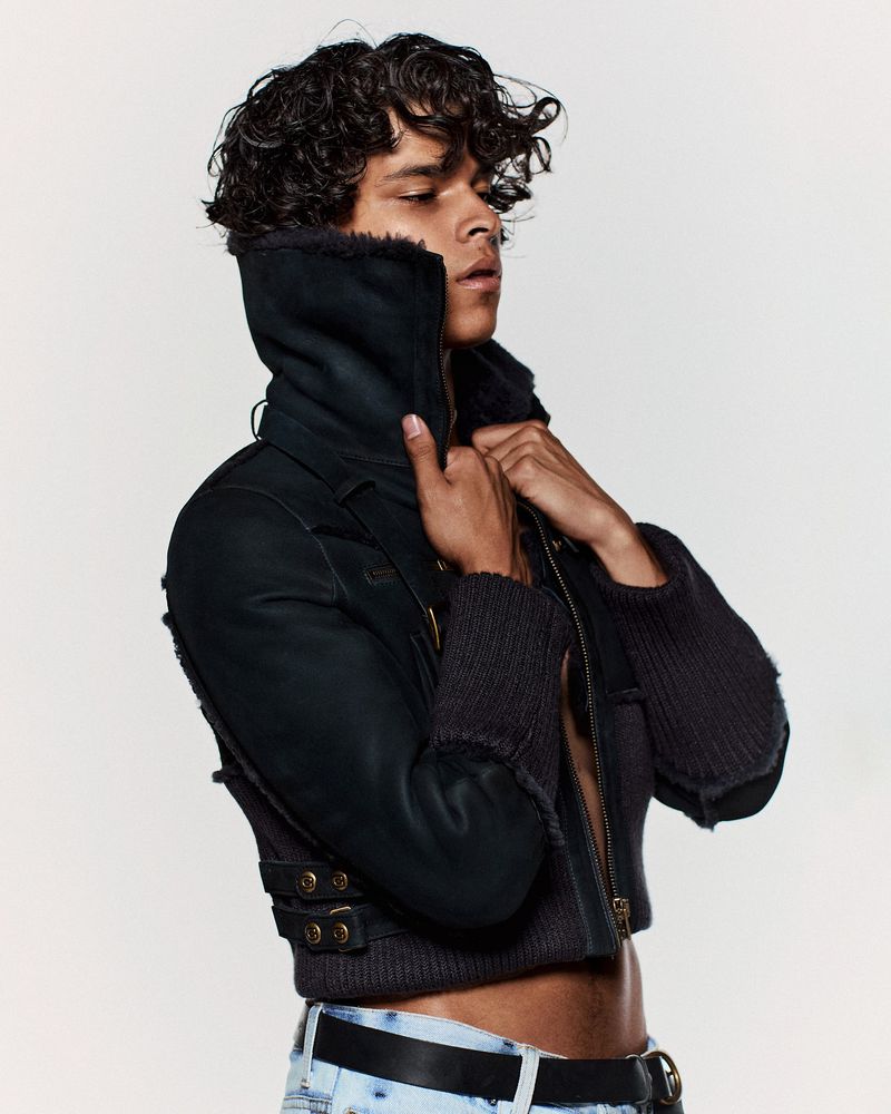 Close-up portrait of a male model with curly hair wearing a black jacket and jeans against a plain white background in a studio setting.
