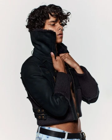 Close-up portrait of a male model with curly hair wearing a black jacket and jeans against a plain white background in a studio setting.