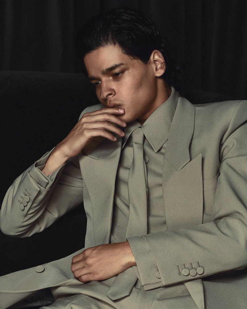Portrait of a man in a sage green suit sitting and looking down with a hand on his face against a dark backdrop. Fashion editorial.