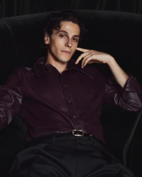 D Pharaoh Woon A Tai seated in a dark velvet armchair wearing a plum button shirt and black trousers for V Man