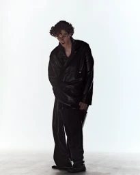 Actor D'Pharaoh Woon-A-Tai standing in an oversized black leather jacket and matching trousers against a bright white backdrop.