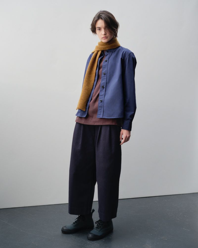 Fashionable adult poses indoors wearing blue collared shirt, brown tee, brown knit scarf, navy pants, and black shoes against plain background.