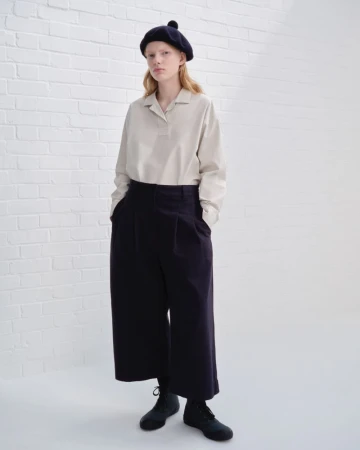 Full-length studio shot of an adult woman in wide-leg pants, long-sleeved shirt, and beret, standing against a white brick wall.