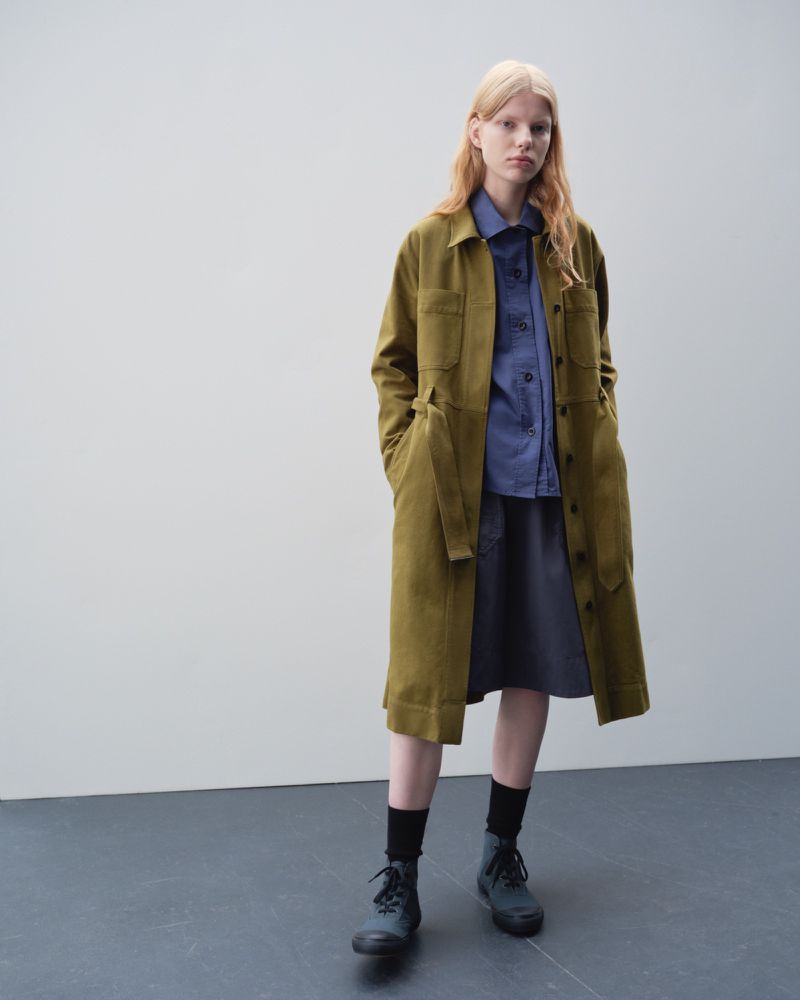 A woman in a long olive jacket, blue shirt, and grey skirt stands in front of a light grey wall in a fashion studio shot.