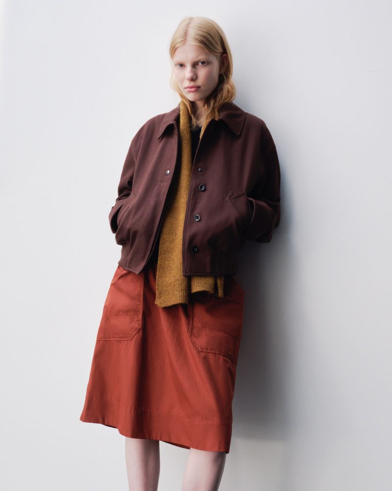 A fair-skinned young woman stands in a studio wearing a brown jacket, gold scarf, and red skirt, showcasing fashion apparel.