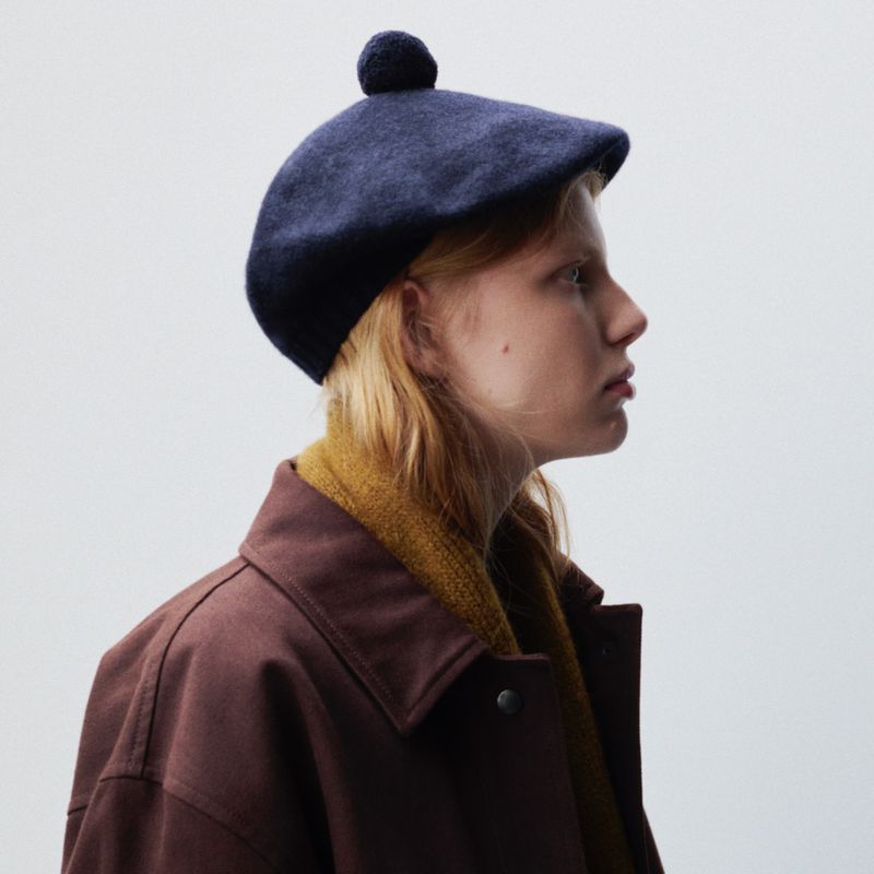 Profile shot of a fair-skinned woman wearing a navy beret with a pompom, a mustard-yellow scarf, and a brown coat against a light background.