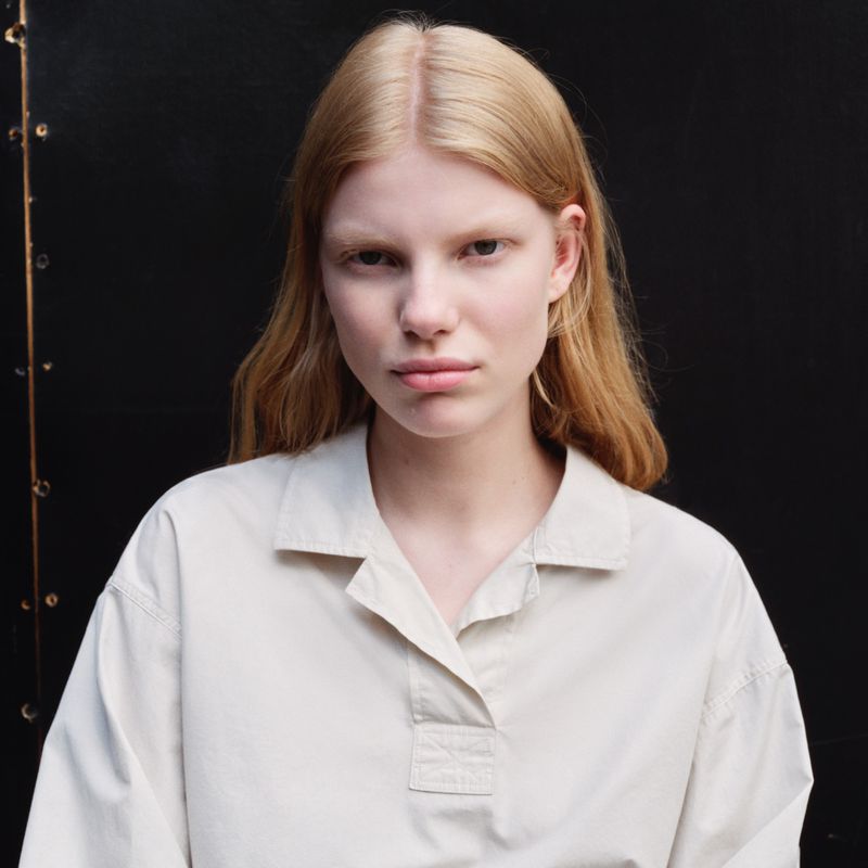 Portrait of a young fair-skinned adult female with blond hair wearing a casual off-white blouse against a dark textured backdrop.
