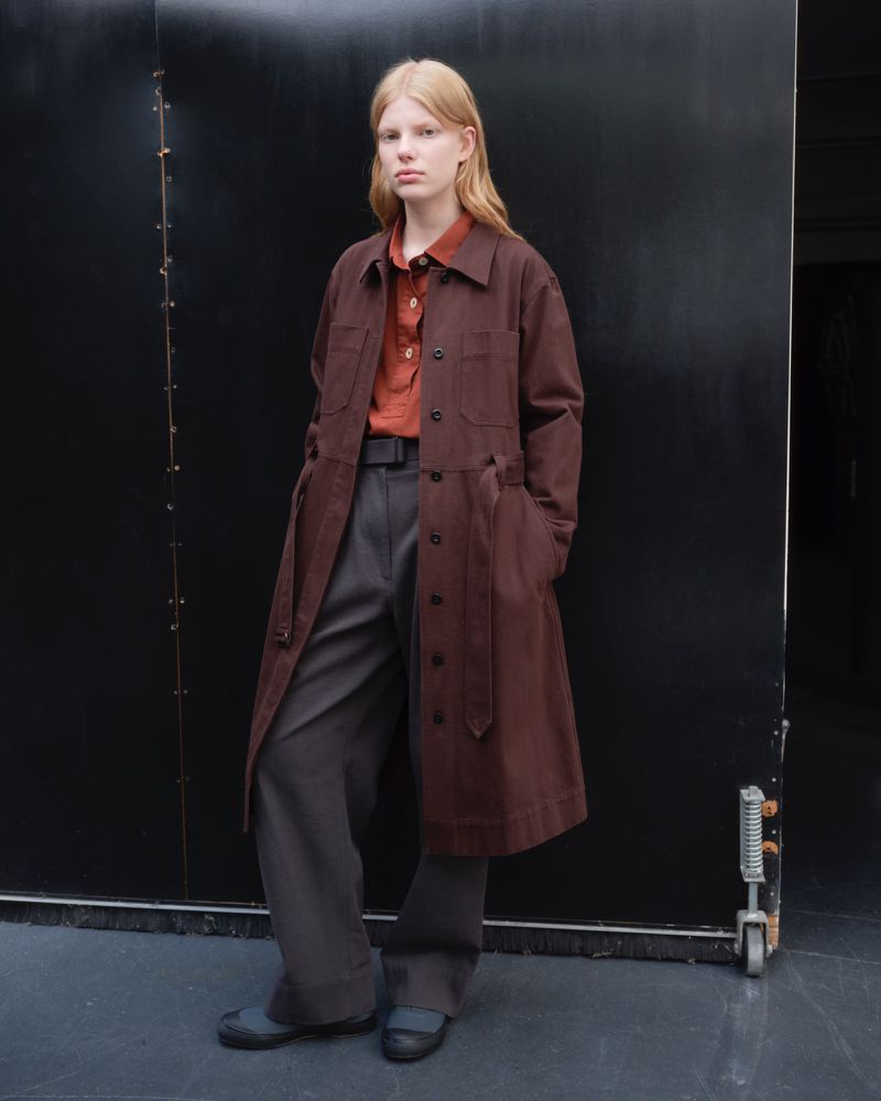 A fair-skinned adult woman with blonde hair poses in a brown overcoat, orange shirt, and gray pants against a dark gray background.