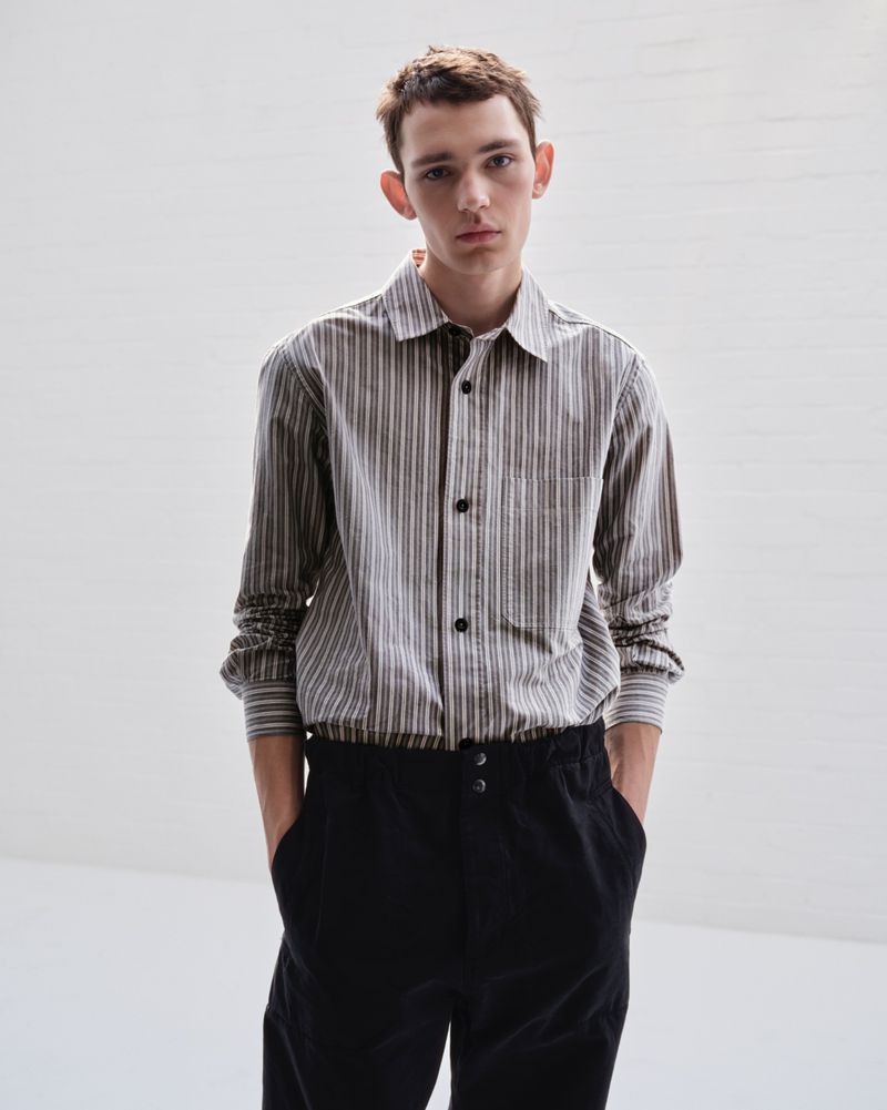 Young adult man in striped long-sleeve shirt and dark pants stands indoors in front of a neutral wall with soft lighting.