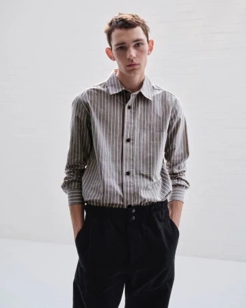 Young adult man in striped long-sleeve shirt and dark pants stands indoors in front of a neutral wall with soft lighting.