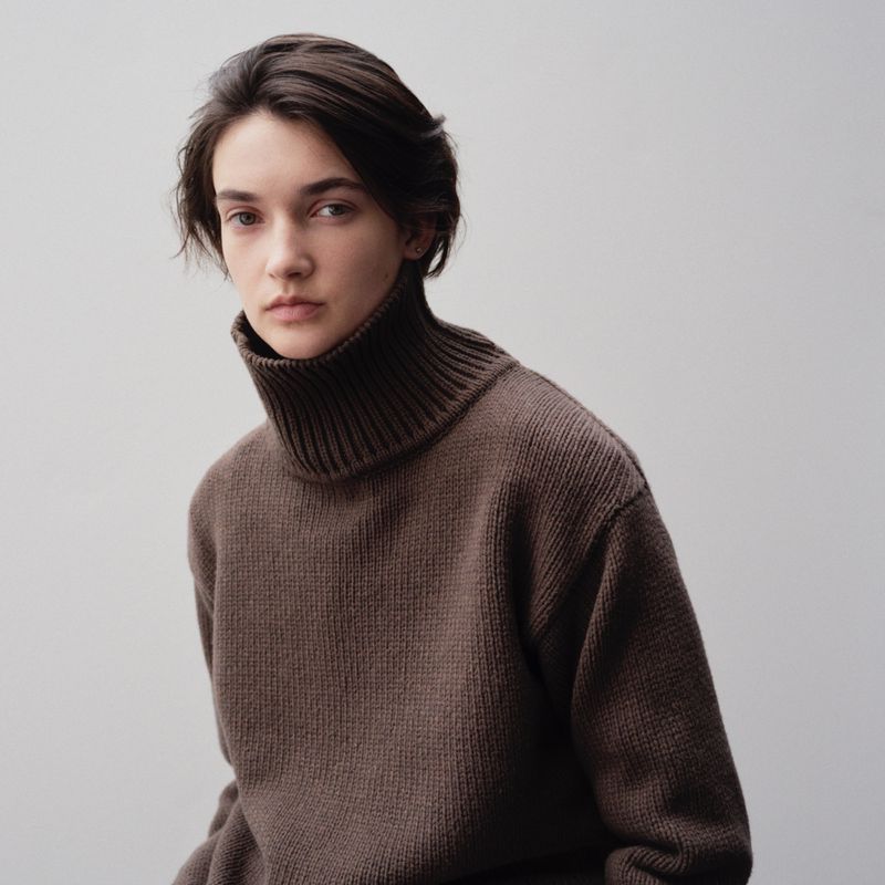 Portrait of an adult woman with dark, short hair wearing a brown turtleneck sweater against a neutral background, looking at the camera.