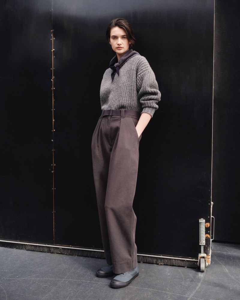 Fashion shot of a woman wearing a gray ribbed sweater, brown trousers, and dark grey shoes standing in front of a black door.