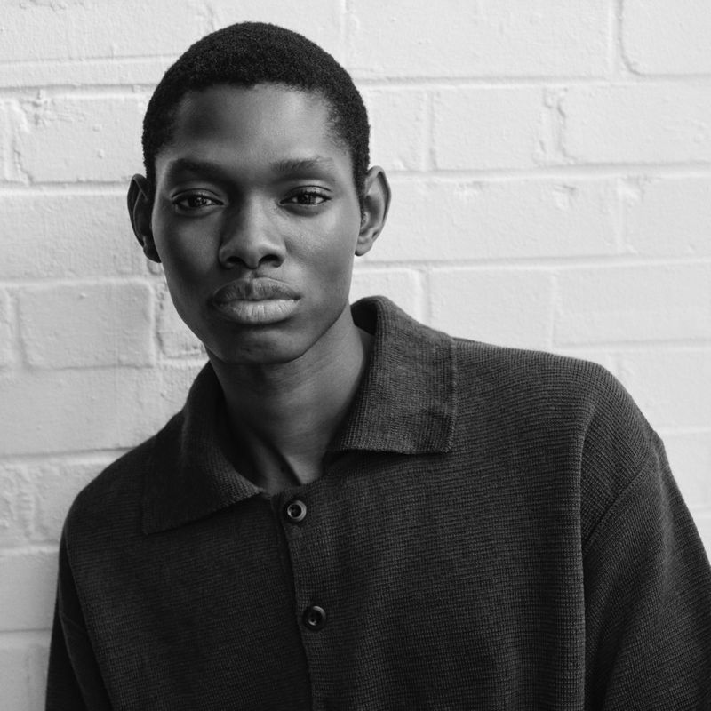 Portrait of a young Black man in a button-down shirt against a white brick wall, captured in black and white with a neutral expression.