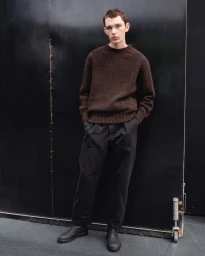Model in a brown knit sweater and dark trousers standing against a black panel background for MHL by Margaret Howell.