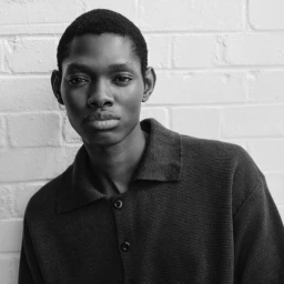Monochrome portrait of model Denetric Malope wearing a dark knit collared shirt against a white painted brick wall.