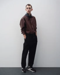 Model standing in a brown button-up shirt and black trousers against a neutral grey studio background.