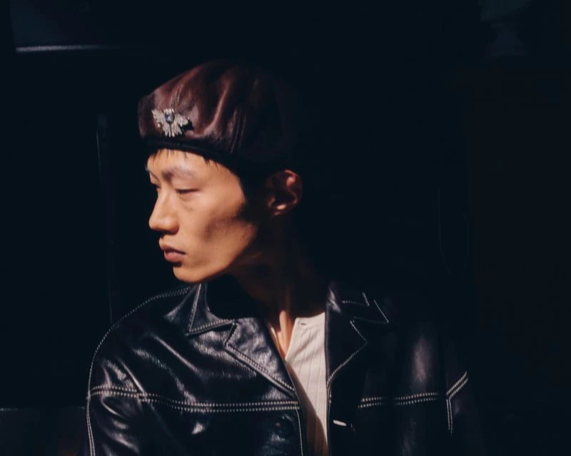 Headshot of an Asian male model wearing a leather beret and jacket. Dramatic chiaroscuro lighting highlights his profile.