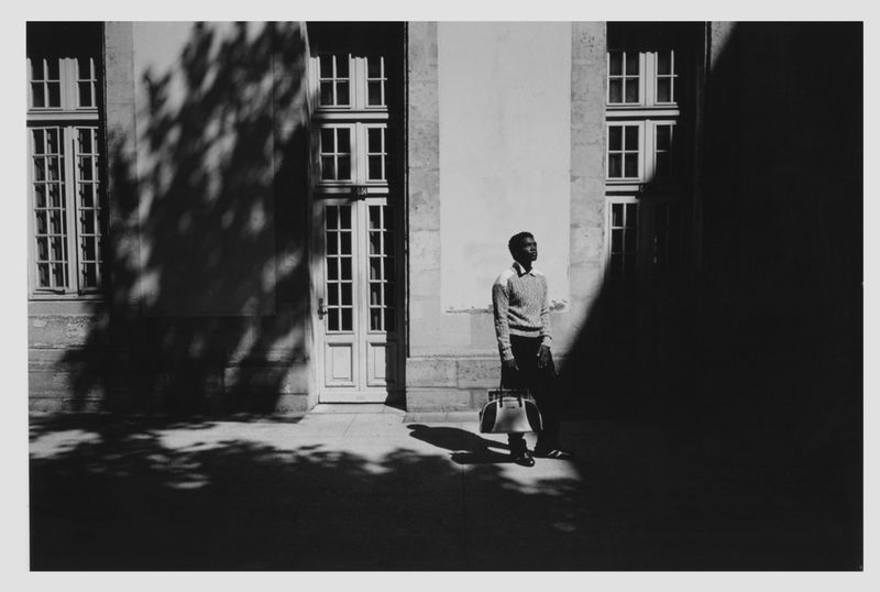 Black and white photograph of a dark-skinned man in a light-colored sweater standing near a building with window and door details.