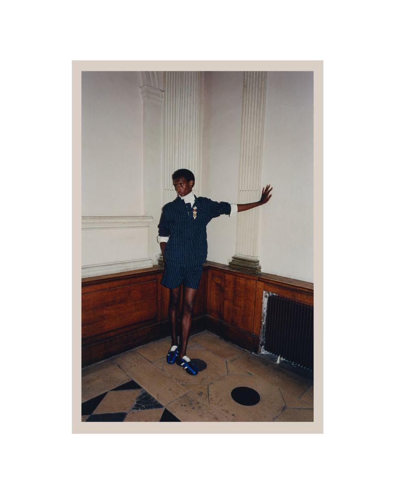 Dark-skinned adult in a pinstripe short suit with arm extended in an interior with marble floor, wood paneling, and fluted columns.