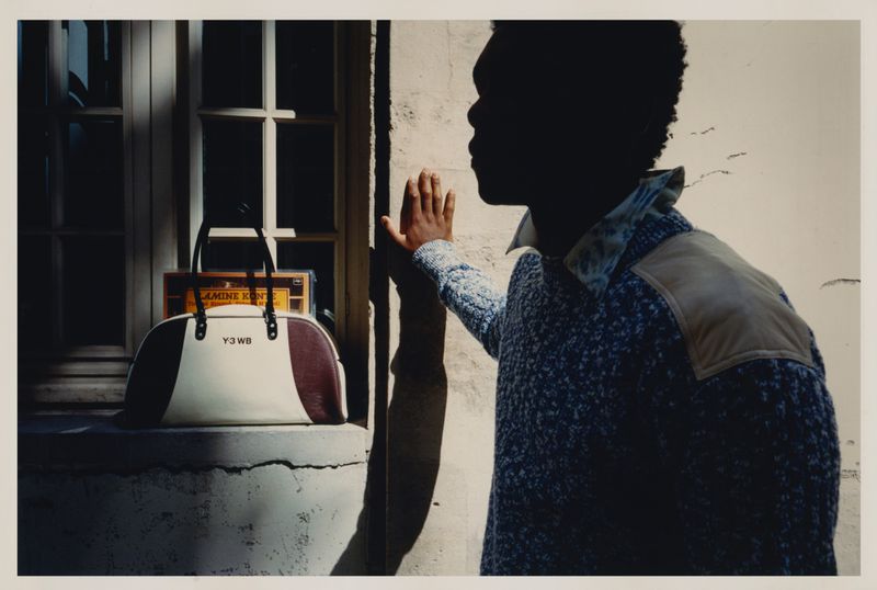 Profile shot of a silhouetted African American male in a blue sweater, next to a beige and burgundy designer bag on a ledge.