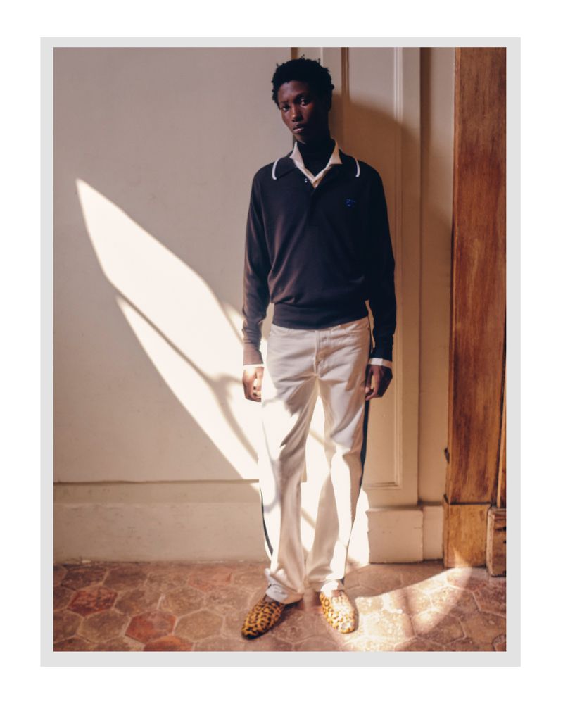 Young Black man in casual-formal fashion stands indoors with sunlight. He wears a dark sweater, white pants, and patterned loafers.