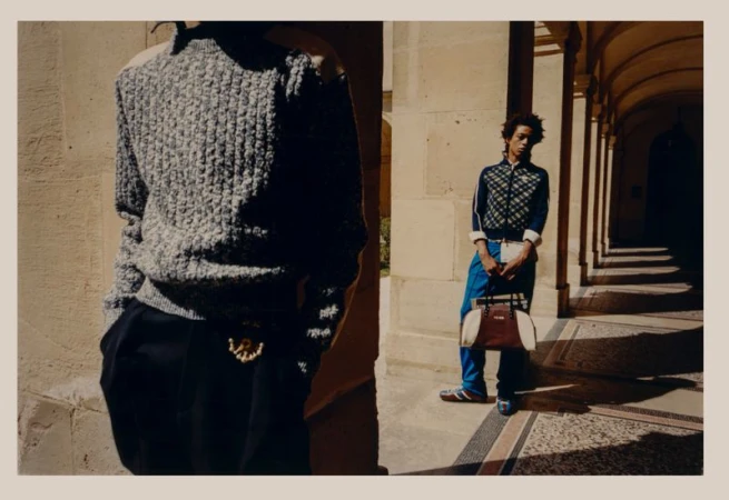 Two models pose in sunlight on a stone colonnade, one in a gray sweater, and the other holding a bag, wearing bright blue pants.