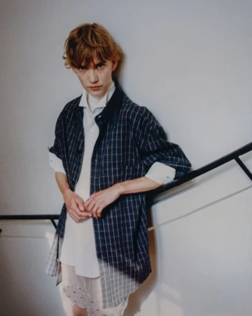 Young adult poses on stairs wearing an unbuttoned navy blue and white plaid shirt over a white top with lace-trimmed sheer bottoms.