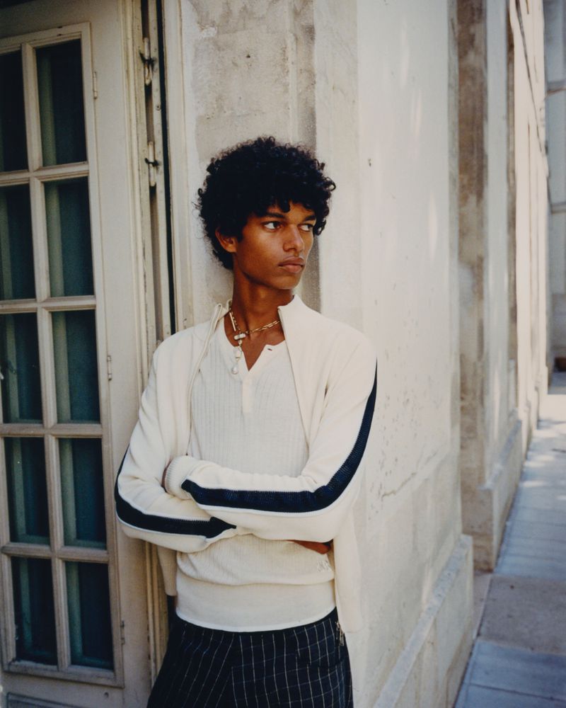 Portrait of a young adult man with dark, curly hair wearing a white shirt and jacket, posing outdoors in front of a stone building.